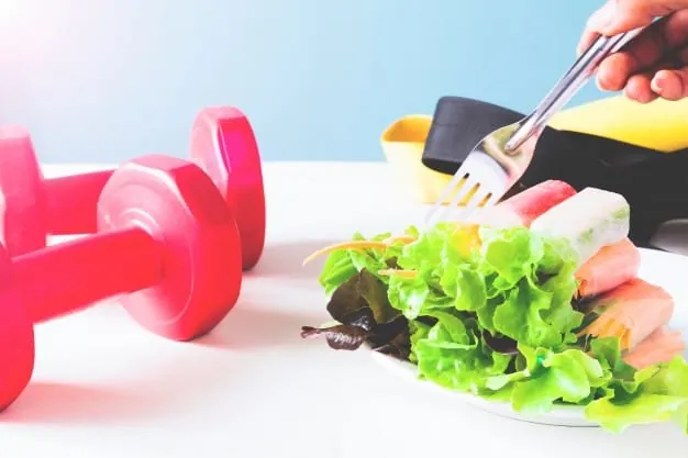 Composición que muestra unas mancuernas rojas junto a un plato de ensalada fresca, representando el equilibrio entre ejercicio físico y alimentación saludable.