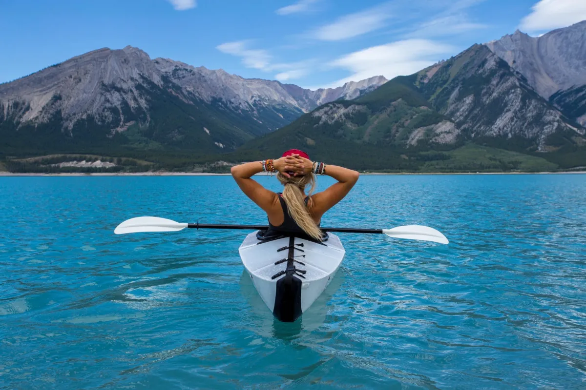 Mujer relajada en un kayak sobre un lago turquesa frente a montañas, representando la unión entre salud y placer.
