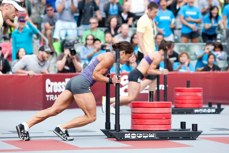 Mujer con musculatura definida empujando un trineo de carga con discos rojos durante una competencia de CrossFit frente a una multitud.