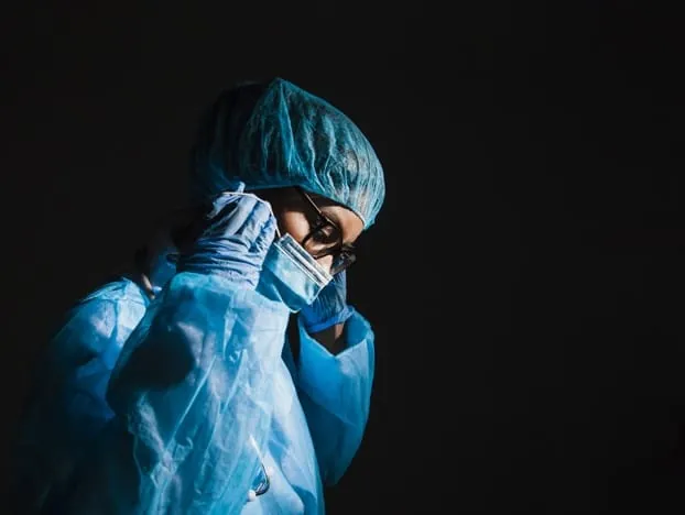 Cirujana con bata azul, mascarilla y gorro quirúrgico ajustándose las gafas en un entorno oscuro