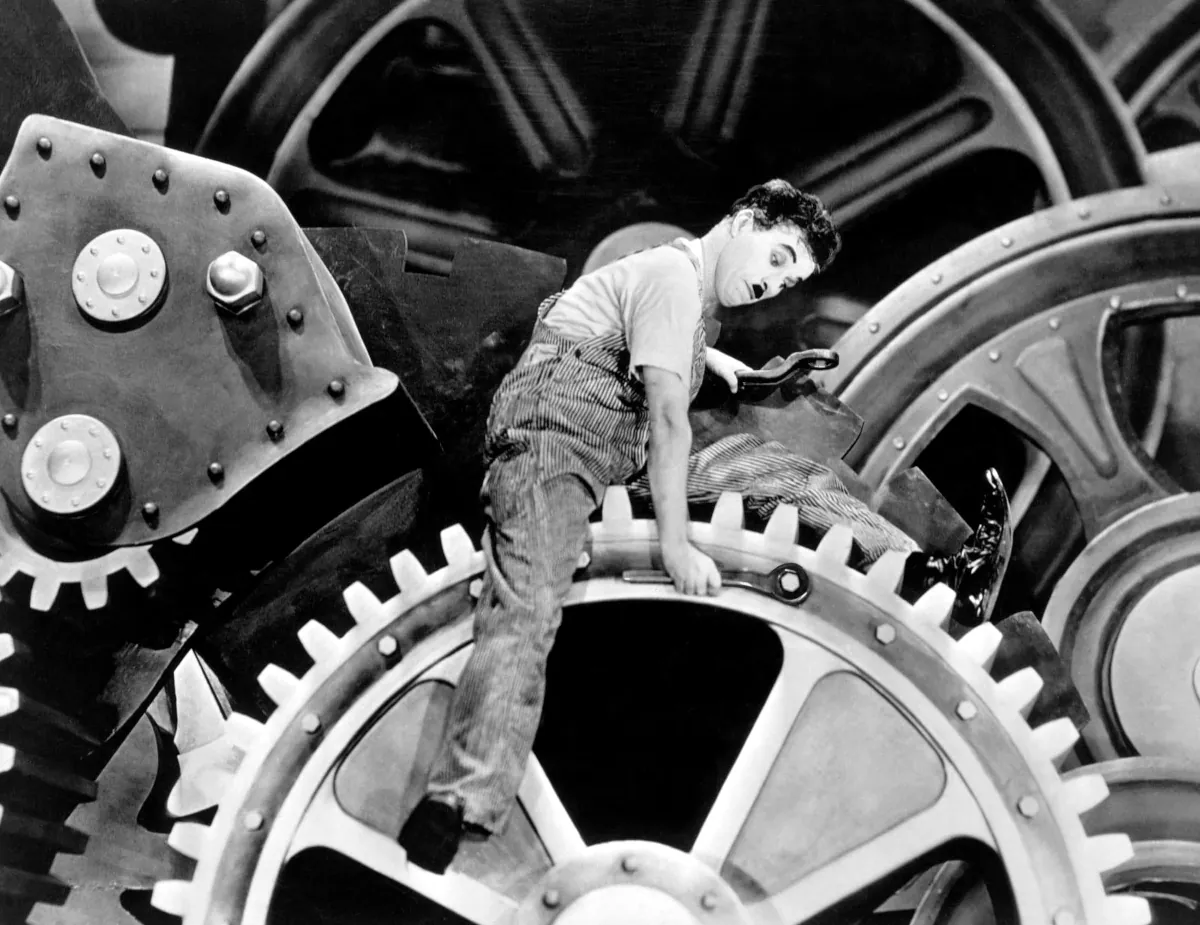 Charles Chaplin atrapado entre engranajes industriales, simbolizando el impacto del trabajo mecanizado en la salud.