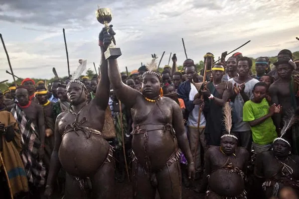 Miembros de la tribu Bodi en Etiopía celebrando el festival Ka'el, mostrando a hombres con abdómenes prominentes y uno de ellos sosteniendo un trofeo, como ejemplo de la influencia del entorno psicosocial en los cánones de salud y belleza.