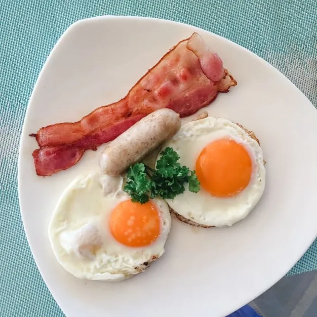 Plato de desayuno compuesto por dos huevos fritos, una salchicha, una tira de bacon y una pequeña guarnición de perejil.