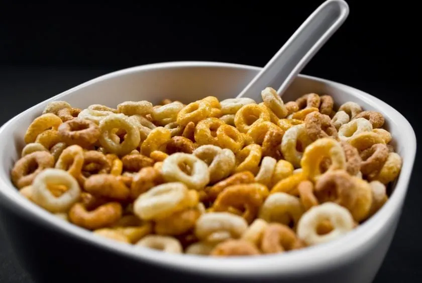 Un cuenco blanco lleno de cereales de desayuno ultraprocesados en forma de aros con una cuchara, sobre un fondo oscuro.