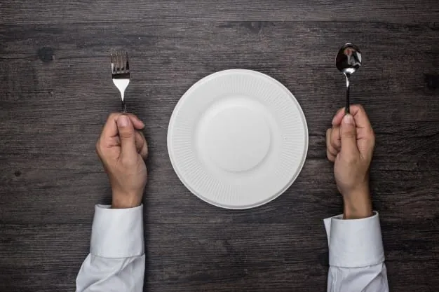 Hambre emocional plato vacio psicologia Manos sosteniendo cubiertos frente a un plato blanco vacío sobre una mesa de madera, representando el concepto de hambre emocional y psicología alimentaria.