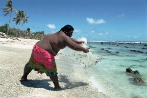 Un hombre en la isla de Nauru lanzando una red de pesca tradicional en la orilla del mar, utilizado como ejemplo de métodos de obtención de alimentos en culturas isleñas y su relación con la alimentación basada en la evolución.