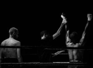 Imagen en blanco y negro de un combate de boxeo donde el árbitro levanta el brazo del ganador en señal de victoria sobre el ring.